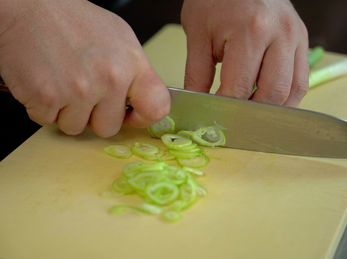 Hands finely chopping fresh green scallions into thin rings with a chefs knife on a yellow cutting board.