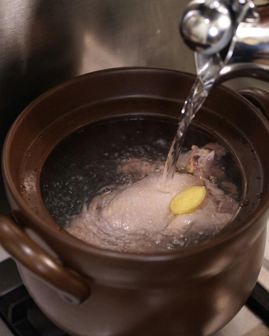Boiling water pouring from a silver kettle over a whole chicken and a ginger slice inside a ceramic pot.
