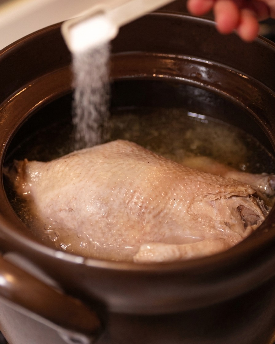 A hand using a small white measuring spoon to sprinkle salt over a whole cooked chicken in clear broth.