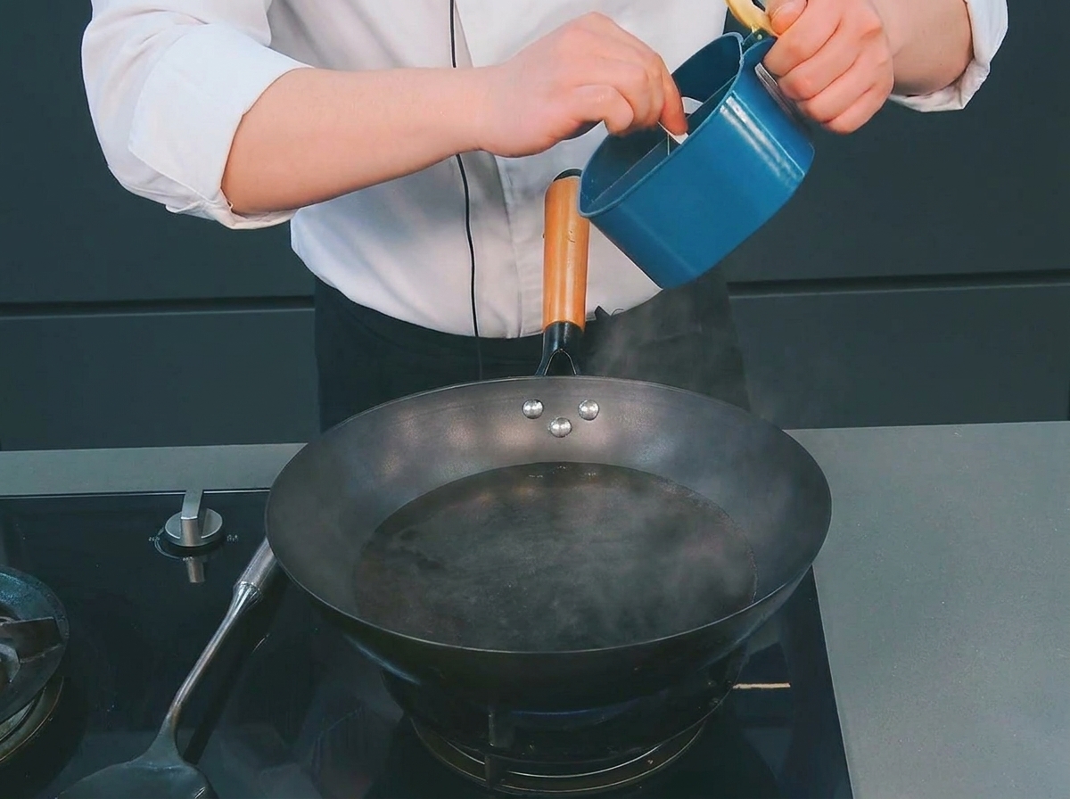 A chef adding a small spoonful of salt from a blue container into a wok of steaming, boiling water.