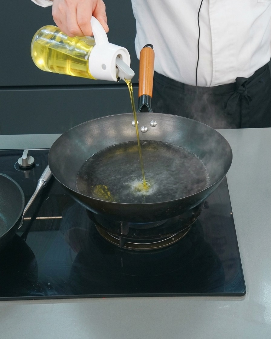 A chef pouring a thin stream of cooking oil from a glass dispenser into a wok of boiling water.