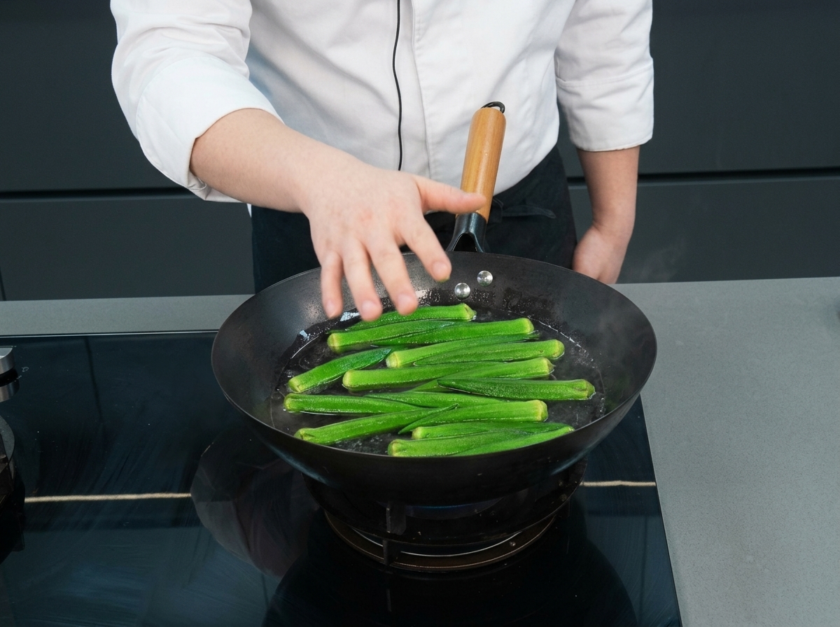 Fresh whole green okra floating in a wok of boiling water as a chefs hand reaches over them.