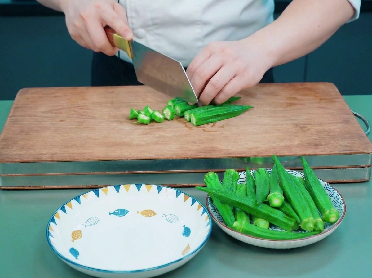 A chef using a cleaver to slice bright green blanched okra into star-shaped rounds on a wooden cutting board.