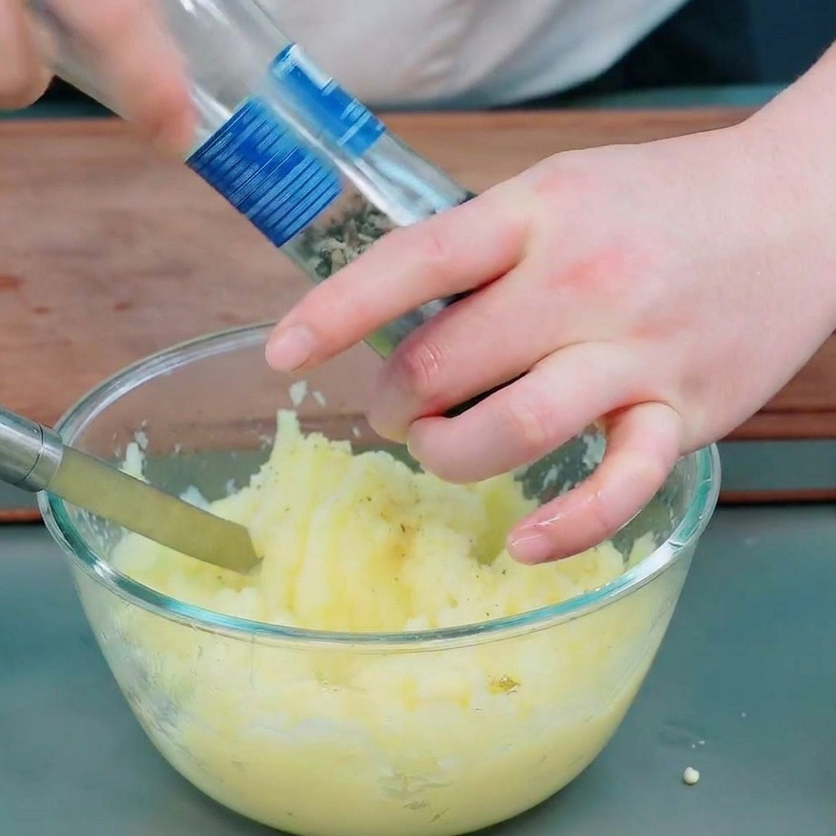 Hands using a glass grinder to add black pepper and salt to smoothly mashed potatoes in a clear glass bowl.