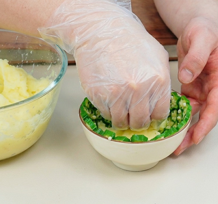 A plastic-gloved hand pressing yellow mashed potatoes into a small white bowl lined with green, star-shaped okra slices.