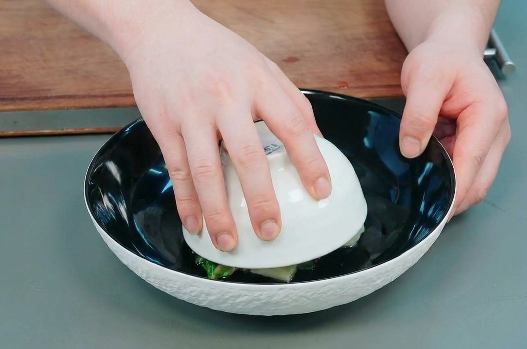Hands carefully lifting a small white bowl away from a large black bowl to reveal the molded okra and potato dome inside.