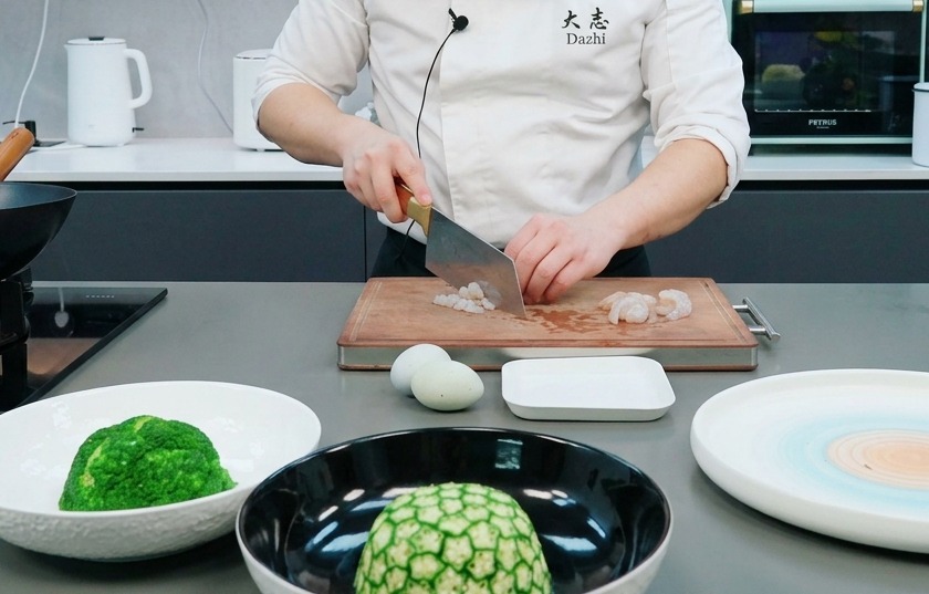 A chef using a large cleaver to chop raw shrimp into small pieces on a wooden cutting board.