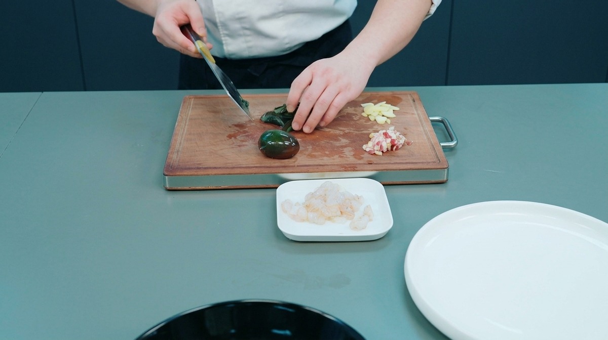 Chef slicing a dark century egg on a wooden cutting board alongside sliced garlic and diced meat.