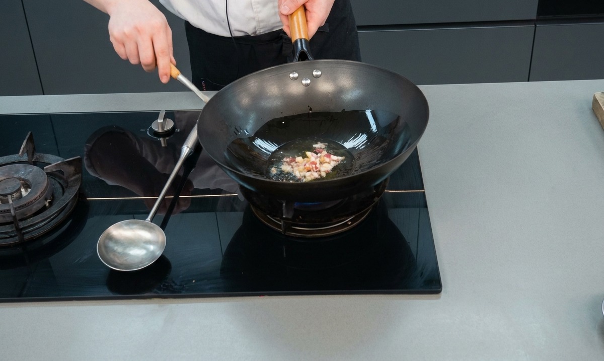 Diced preserved meat and garlic sizzling in hot oil inside a black wok over a gas stove.