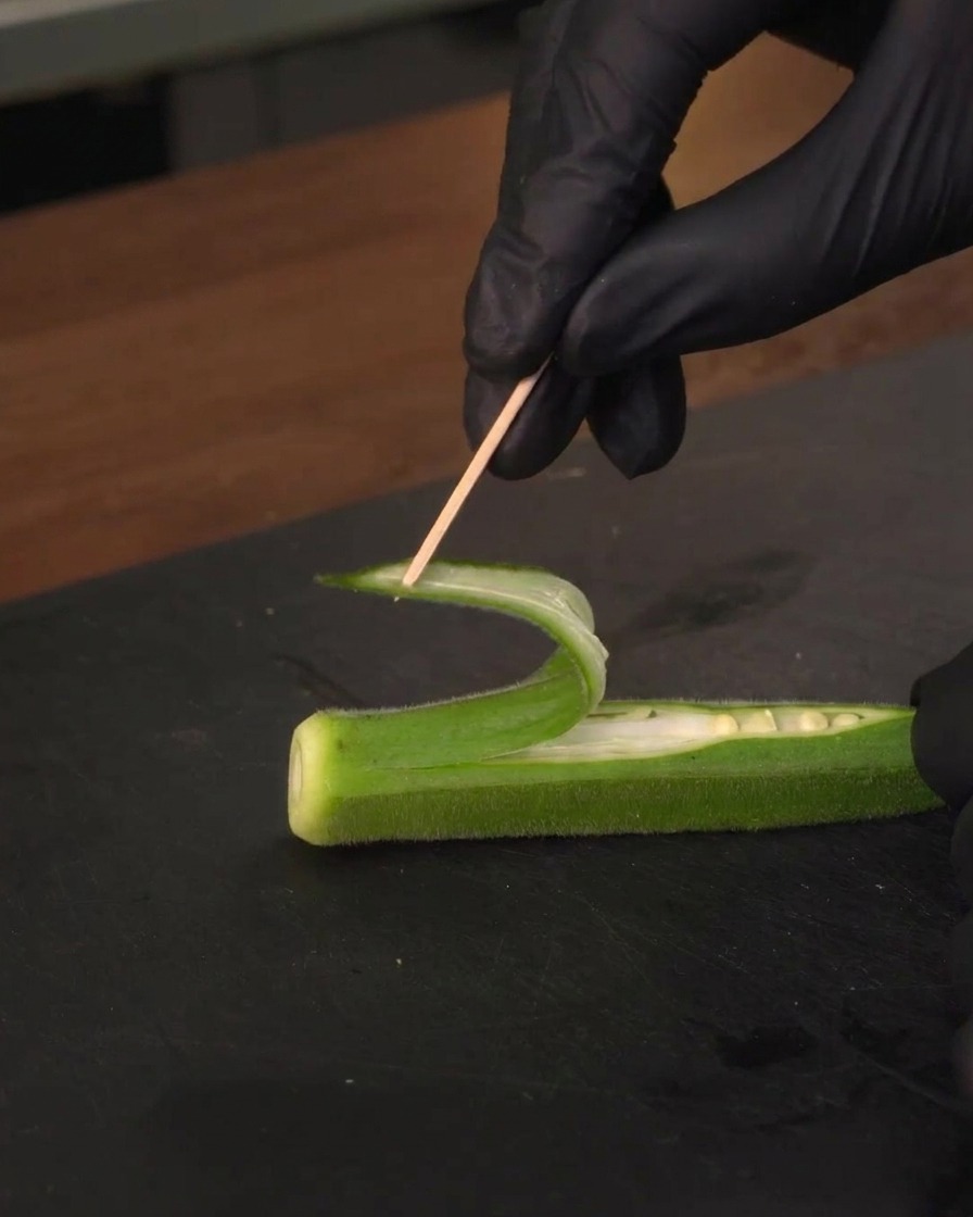 Pinning a curled strip of okra backward using a wooden toothpick to form a sail.