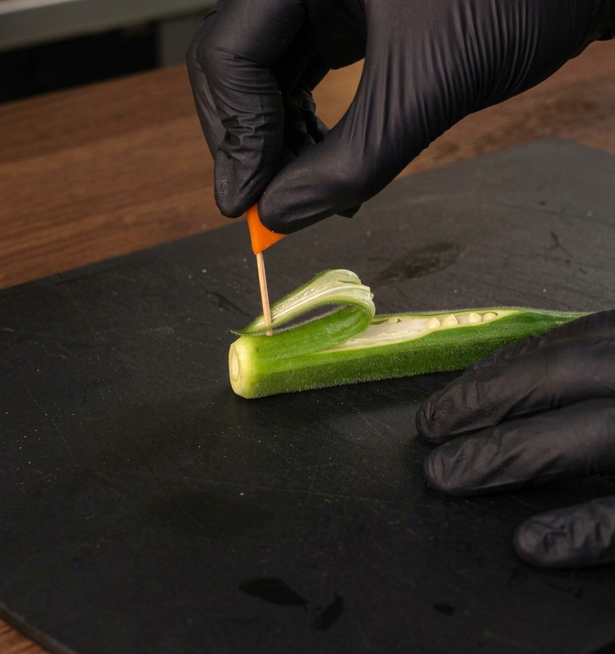 A gloved hand inserting a small orange flag on a toothpick into the okra sail.