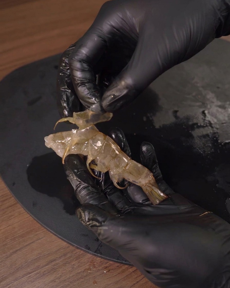 A person wearing black gloves peeling the translucent shell off a raw shrimp.