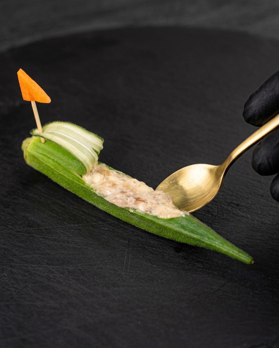 A small golden spoon filling a hollowed-out piece of okra with raw shrimp paste, decorated with a small carrot flag on a toothpick.