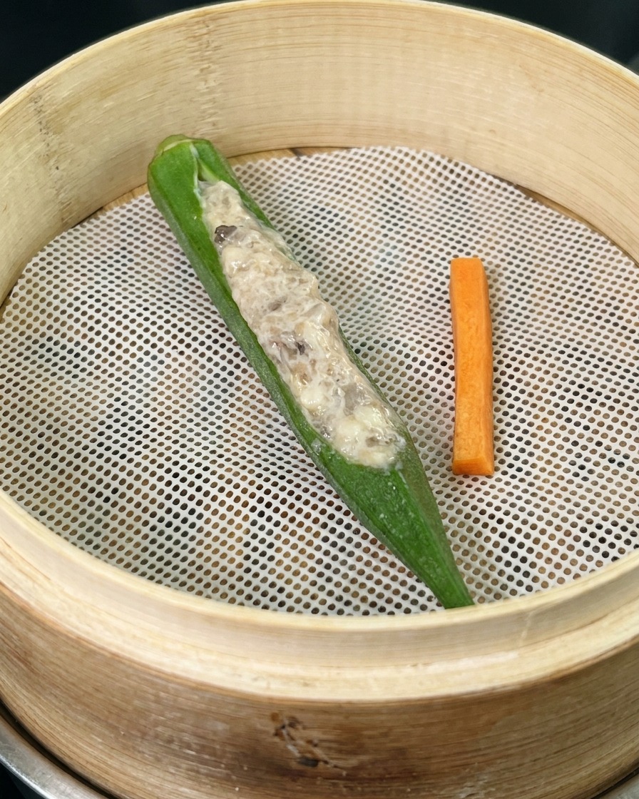 A single meat-stuffed okra pod and a small carrot stick resting on a white mesh liner inside a bamboo steamer basket.