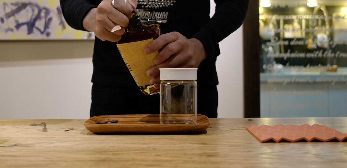 Hands pouring brown bourbon from a bottle into a small, clear glass jar on a wooden surface.