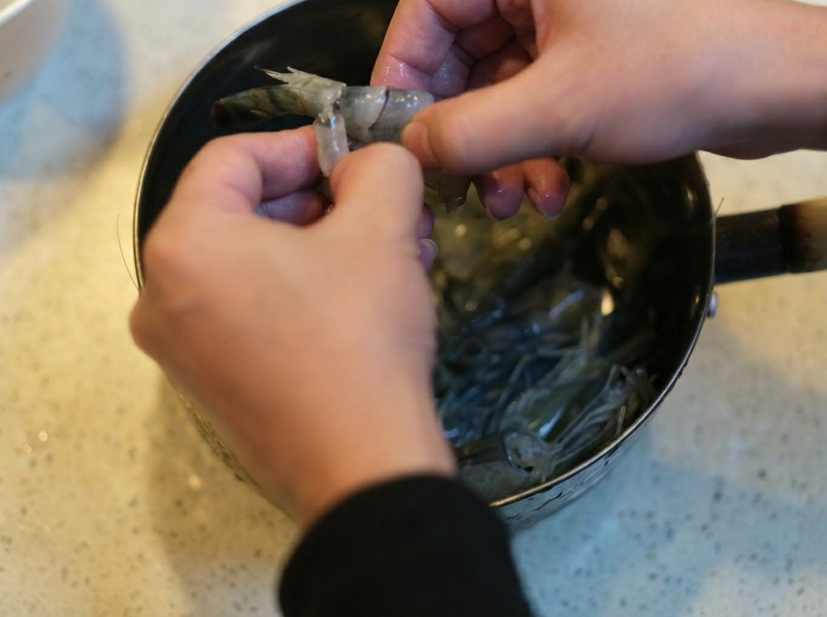 Two hands carefully peeling and deveining raw black tiger shrimp over a metal pot.