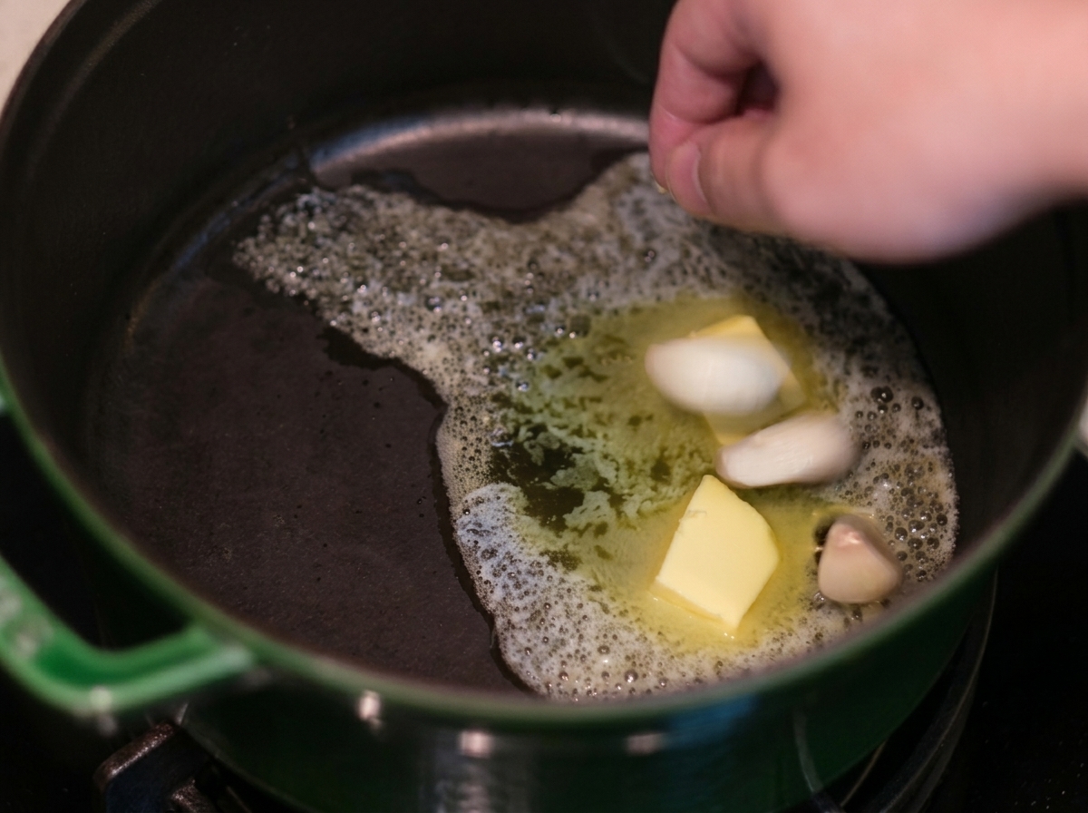 A hand dropping garlic cloves into foaming melted butter inside a green cast-iron pot.