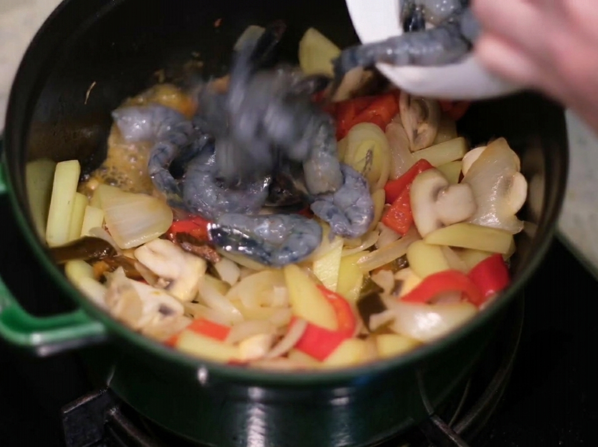 Adding raw, peeled grey shrimp into a green cast-iron pot with sautéed onions, peppers, and potatoes.