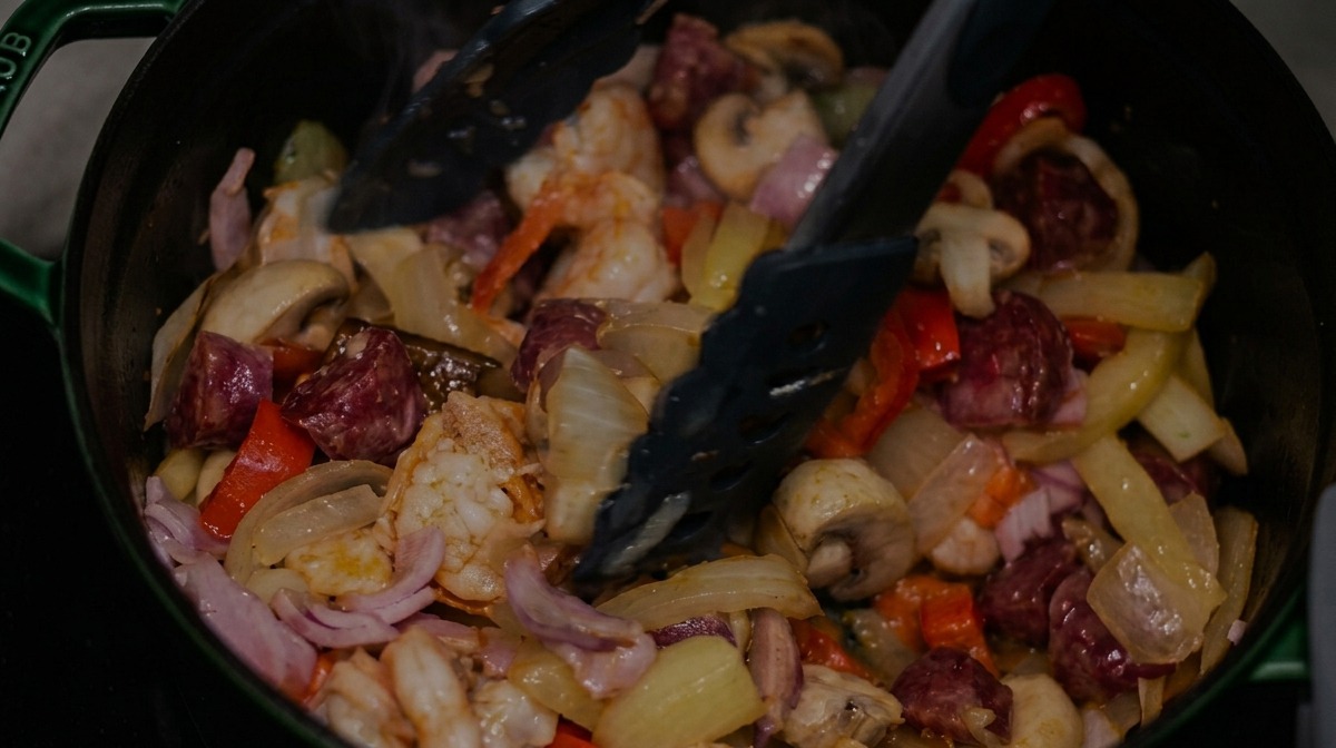 Stirring sliced smoked sausage, bacon, and shrimp into a pot of mixed vegetables with black tongs.