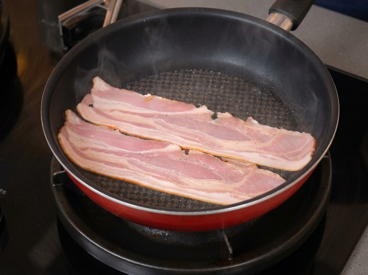 Two long slices of bacon sizzling and starting to curl in a red non-stick frying pan.