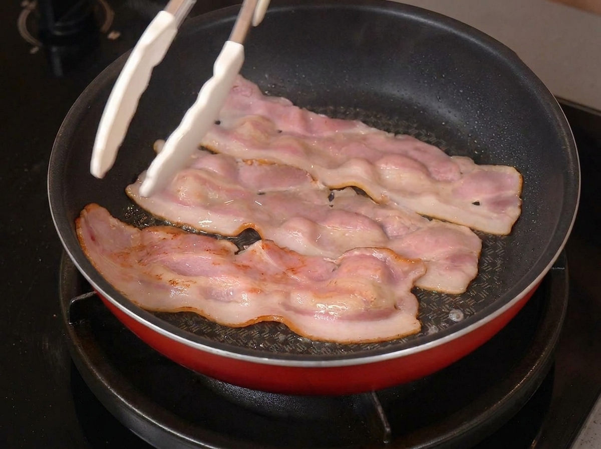 Three strips of bacon sizzling in a frying pan, their edges browning and curling, with a pair of white tongs reaching in.