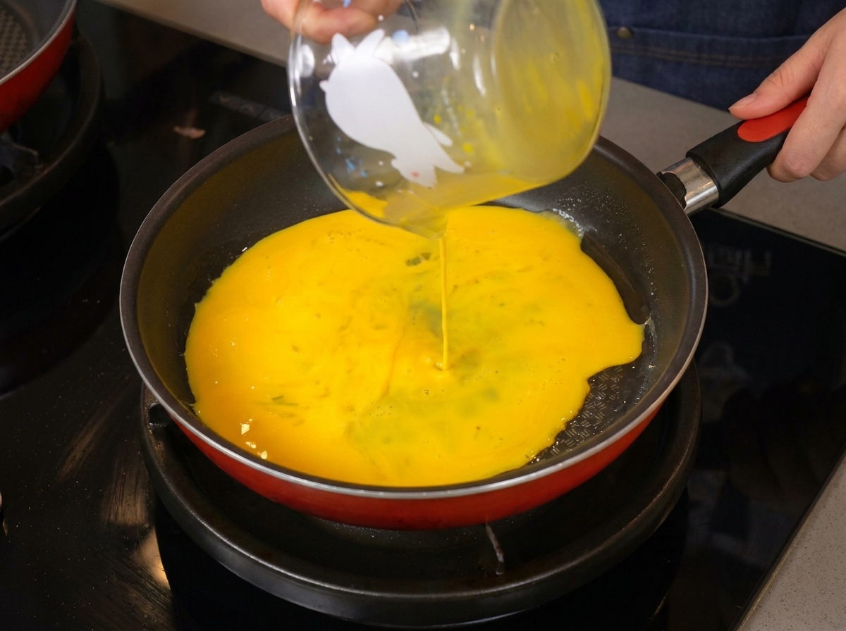 Smooth, bright yellow beaten eggs being poured from a glass measuring cup into a hot frying pan.