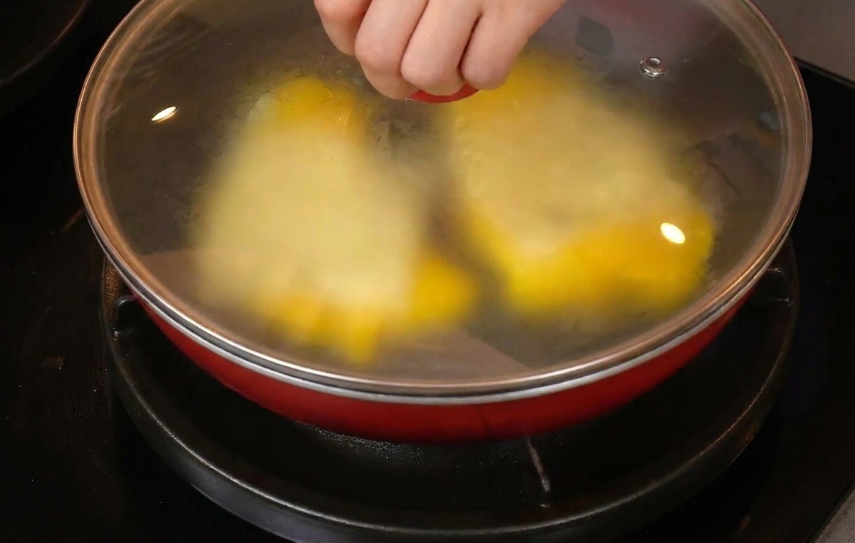 A hand placing a glass lid onto a red frying pan containing scrambled eggs topped with slices of white cheese.