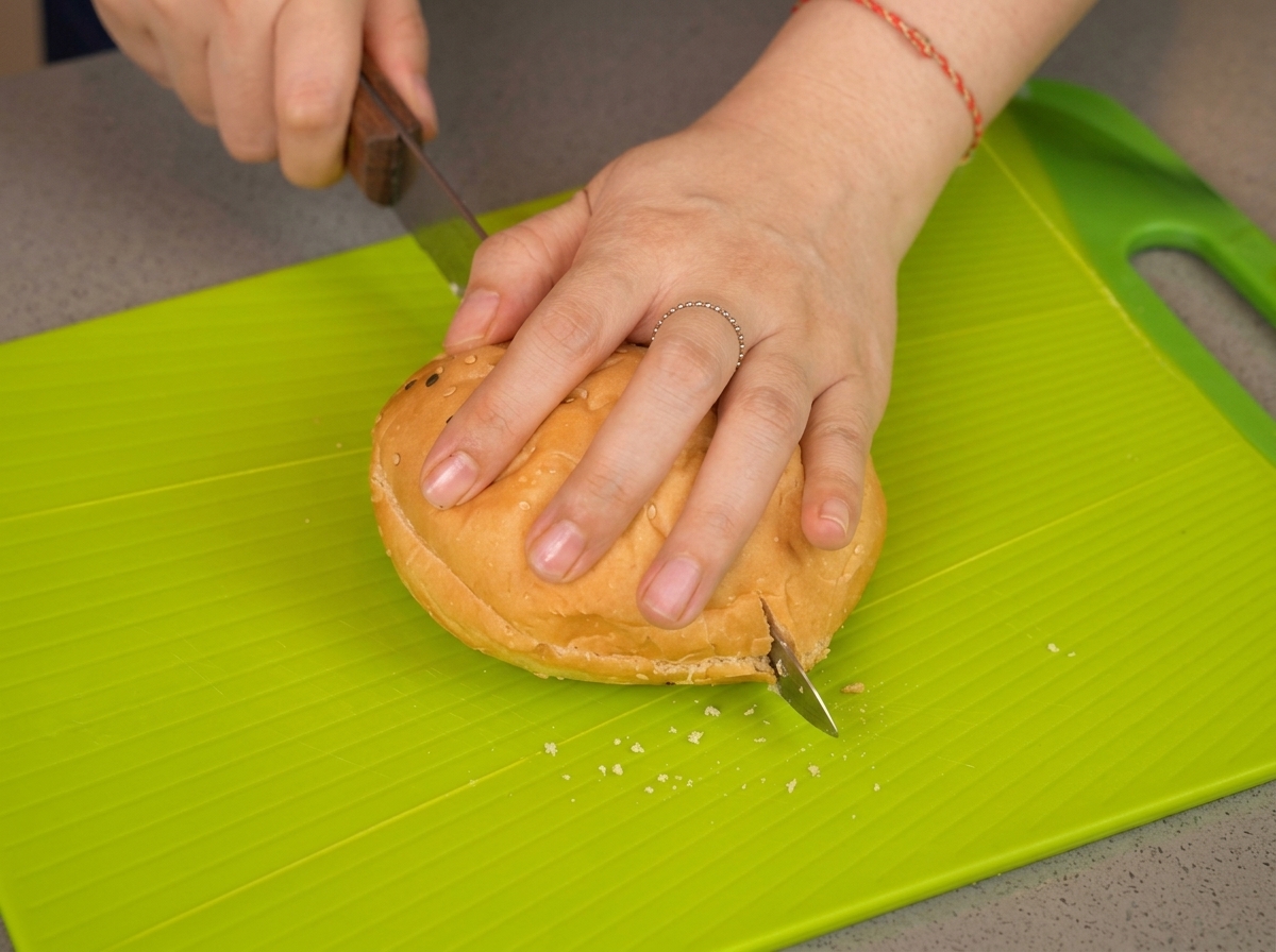 A persons hand pressing down on a sesame bun while a knife slices through the middle on a lime green cutting board.