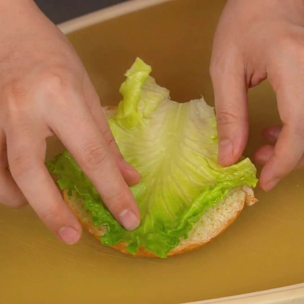 Hands placing a vibrant green lettuce leaf onto the cut surface of a burger bun base.