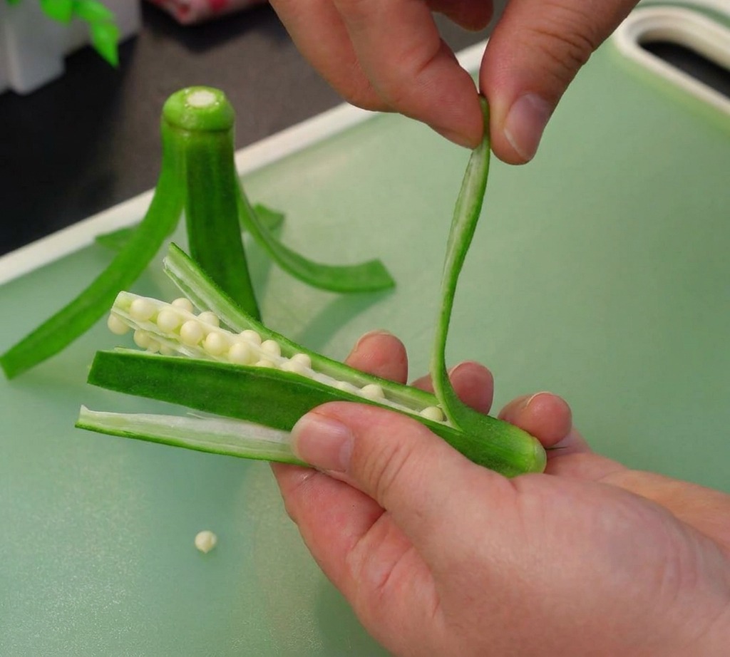 Hands gently separating the outer strips of a scored okra pod to reveal and remove the white seed core.