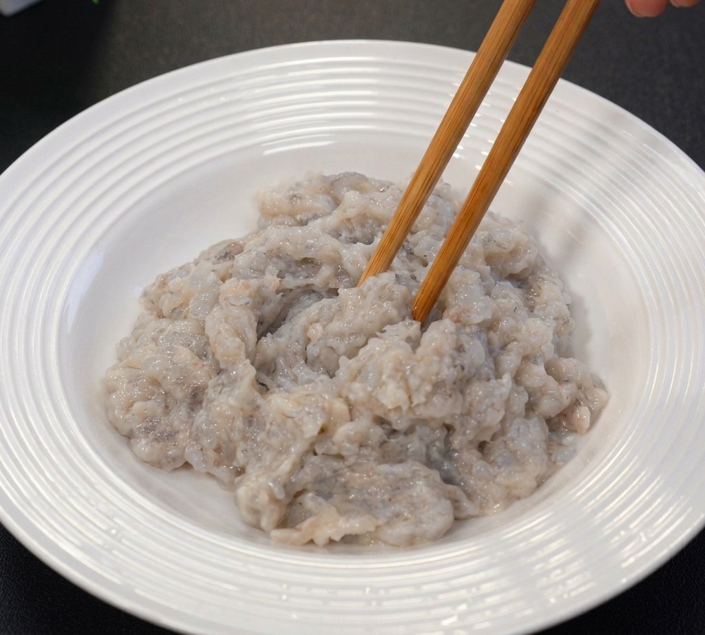 Mixing grayish minced raw shrimp paste in a white ceramic bowl using wooden chopsticks.