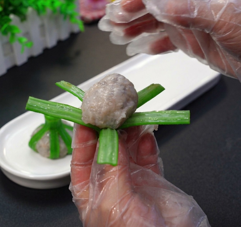 Gloved hands carefully shaping raw shrimp paste into a ball and wrapping it inside the green strips of a blanched okra.