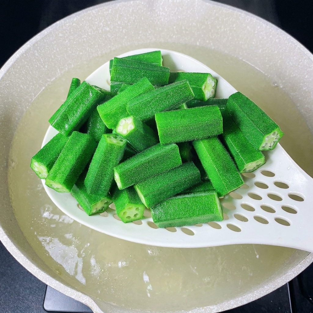 A white slotted spoon lifting freshly blanched okra pieces out of boiling water.