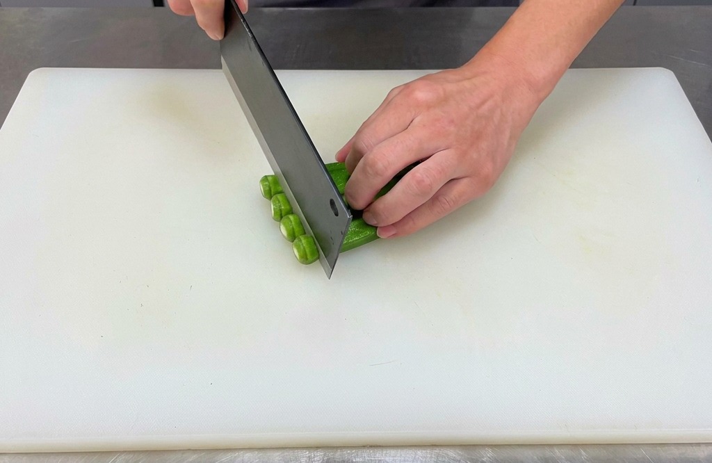 A cook using a cleaver to slice the ends off a row of fresh green okra on a white cutting board.