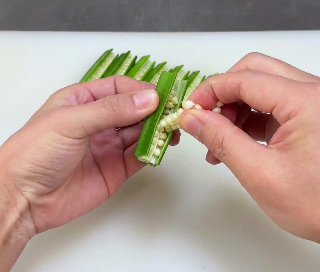 Hands holding a halved piece of green okra and carefully peeling out the white seeds from the center.