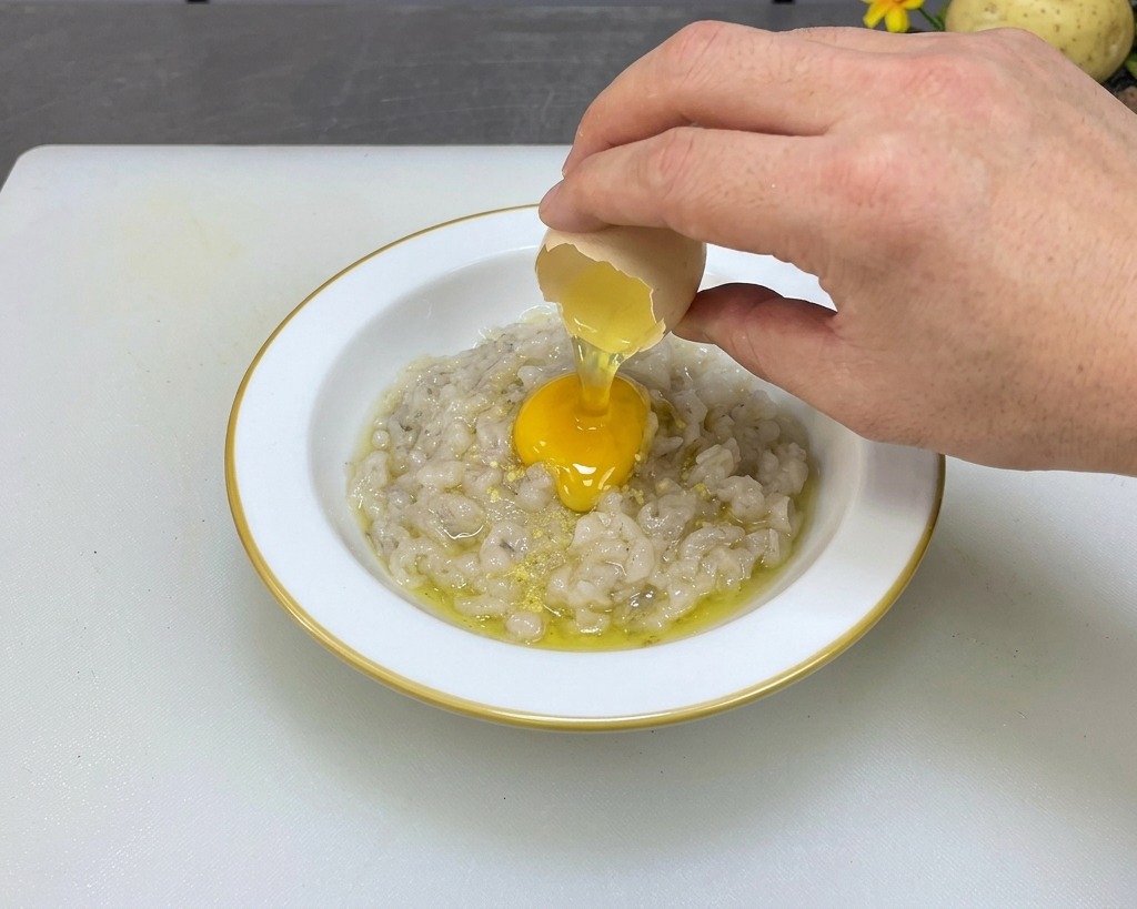 A hand cracking a fresh egg over a bowl of seasoned gray shrimp paste.