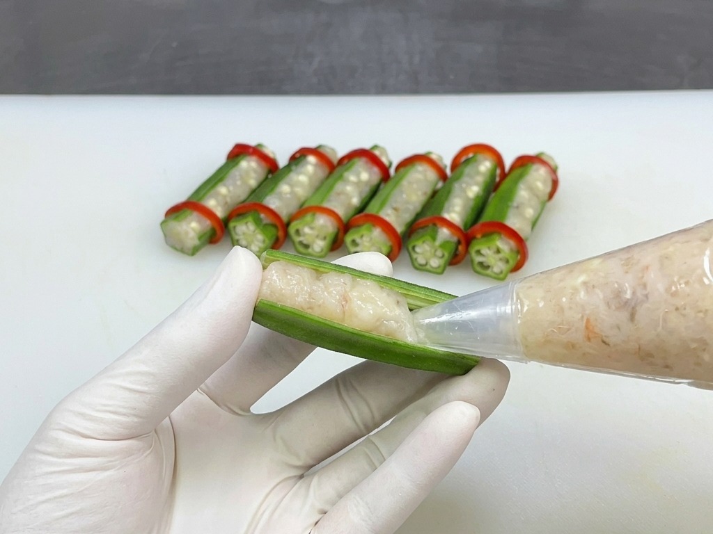 A gloved hand using a clear piping bag to squeeze seasoned shrimp paste into a hollowed green okra half.