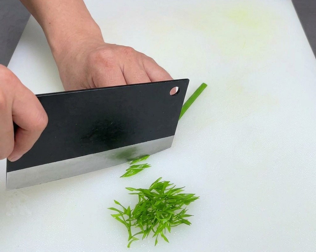Hands using a large black cleaver to thinly slice green scallions on a white cutting board.