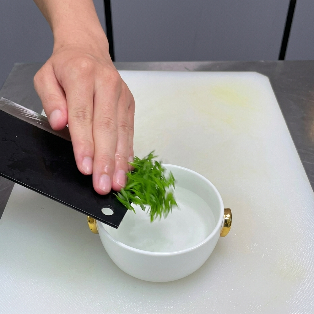 Sliced green scallions being transferred from a black cleaver into a small white bowl of water.