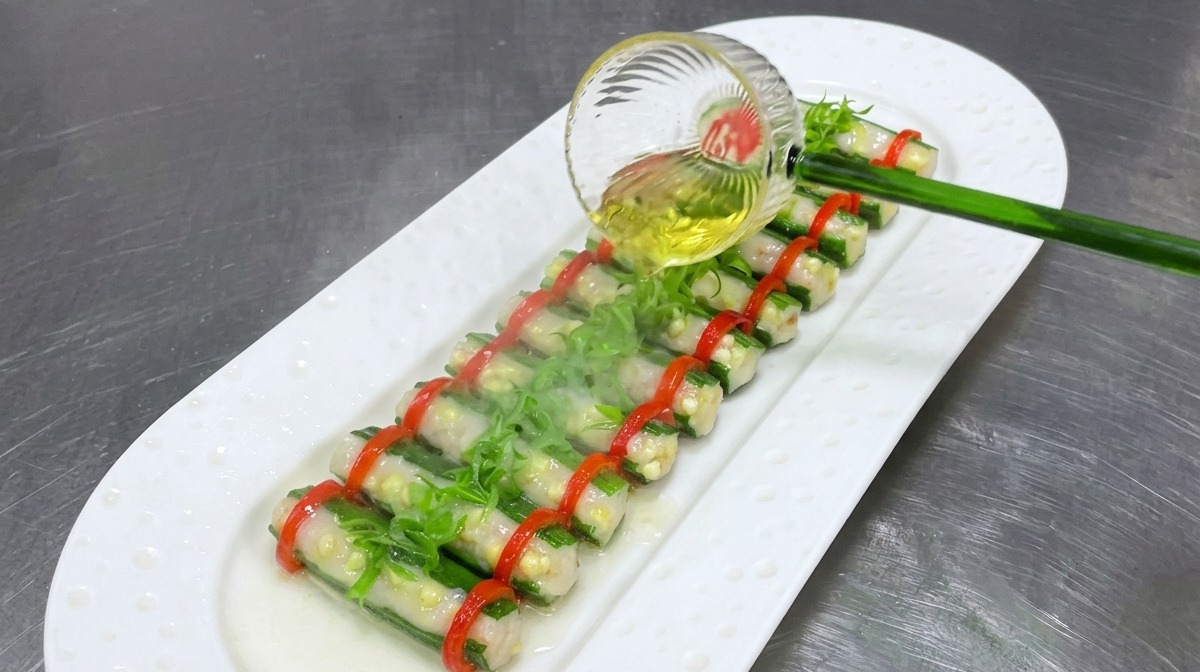 Clear hot oil being poured from a glass ladle over a plate of steamed stuffed okra topped with fresh green scallions and red chili rings.
