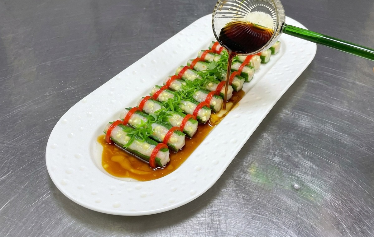 A small glass ladle drizzling a dark savory soy sauce dressing over a beautifully arranged plate of steamed stuffed okra.
