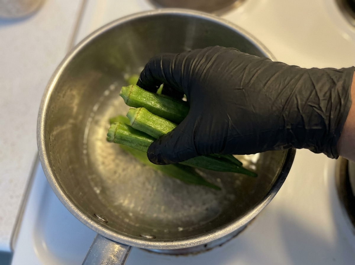 A gloved hand placing fresh whole green okra pods into a metal pot filled with boiling water.