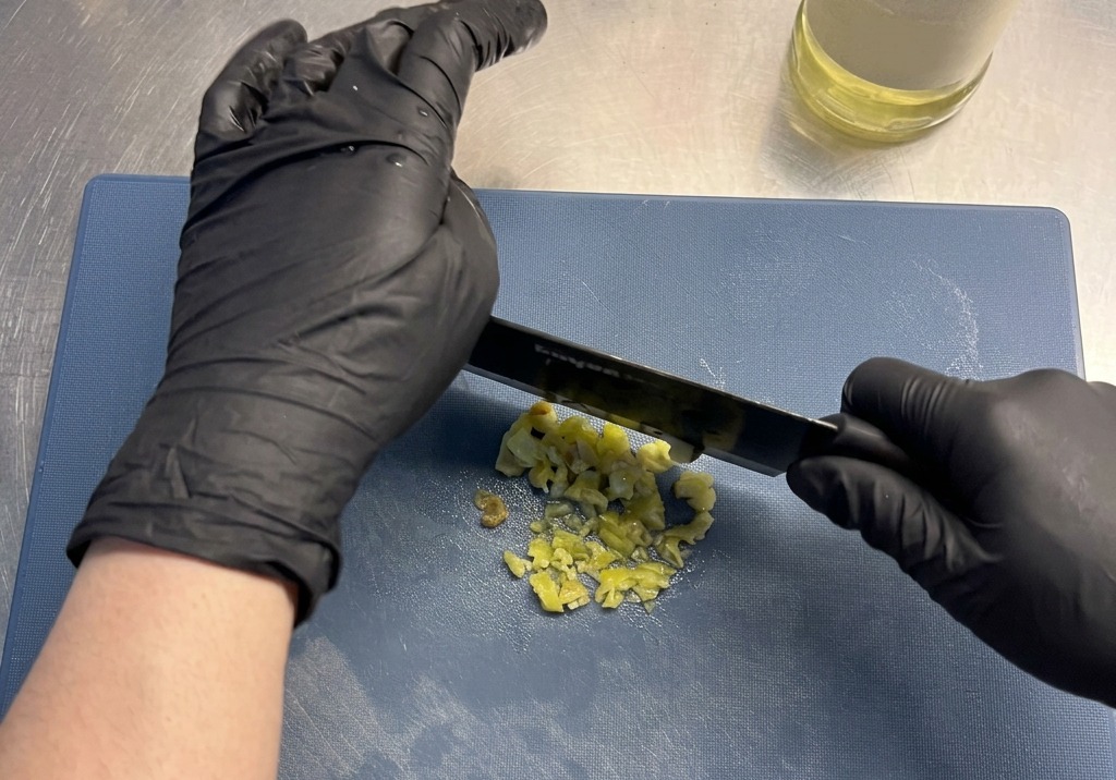 Gloved hands using a black kitchen knife to finely chop yellow pickled peppers on a blue cutting board.
