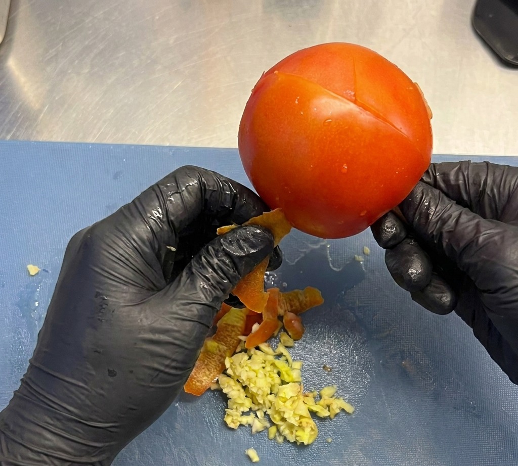 Gloved hands gently peeling the red skin off a whole plump tomato over a cutting board.