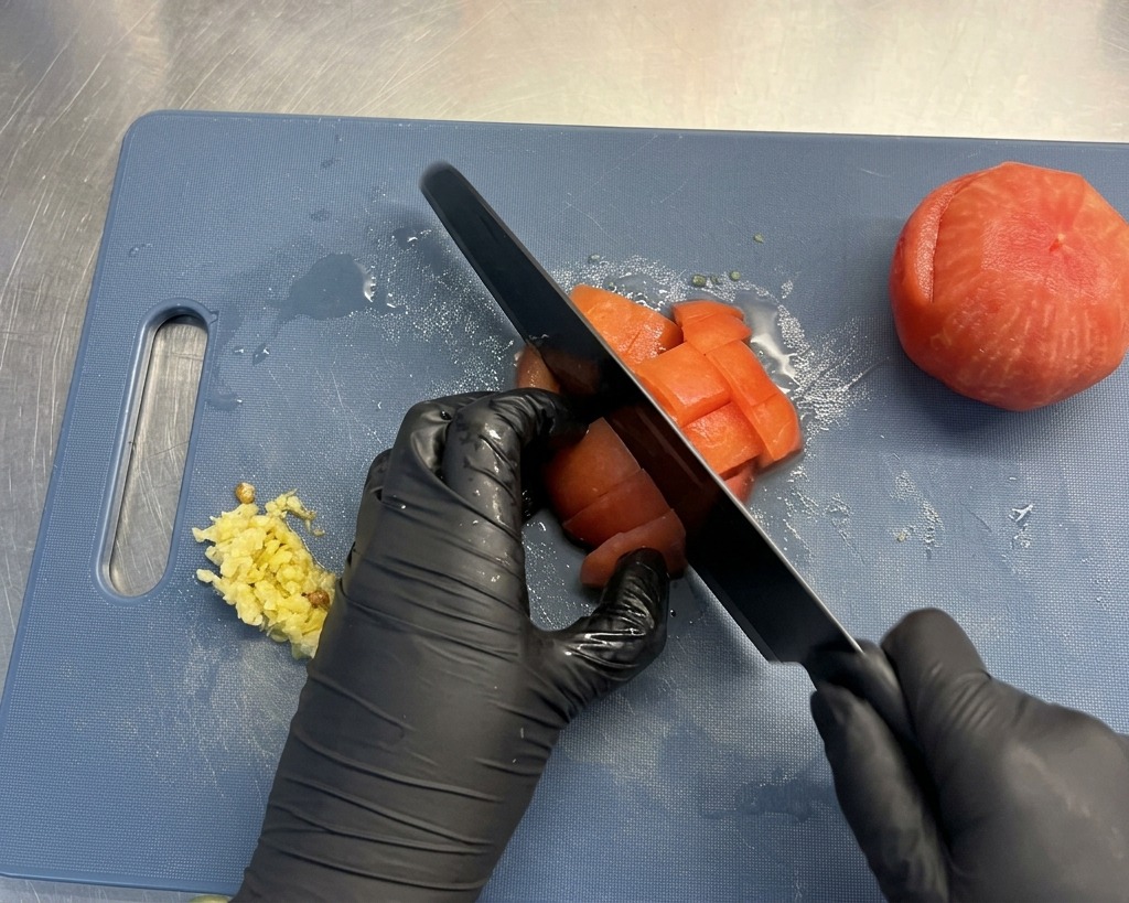 Gloved hands holding a black knife, dicing a peeled red tomato into cubes on a blue cutting board.