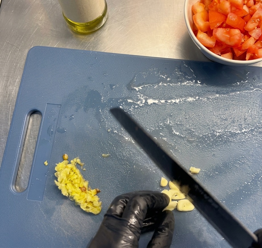 Gloved hands slicing fresh garlic cloves into thin slivers on a blue cutting board, with a bowl of diced tomatoes nearby.