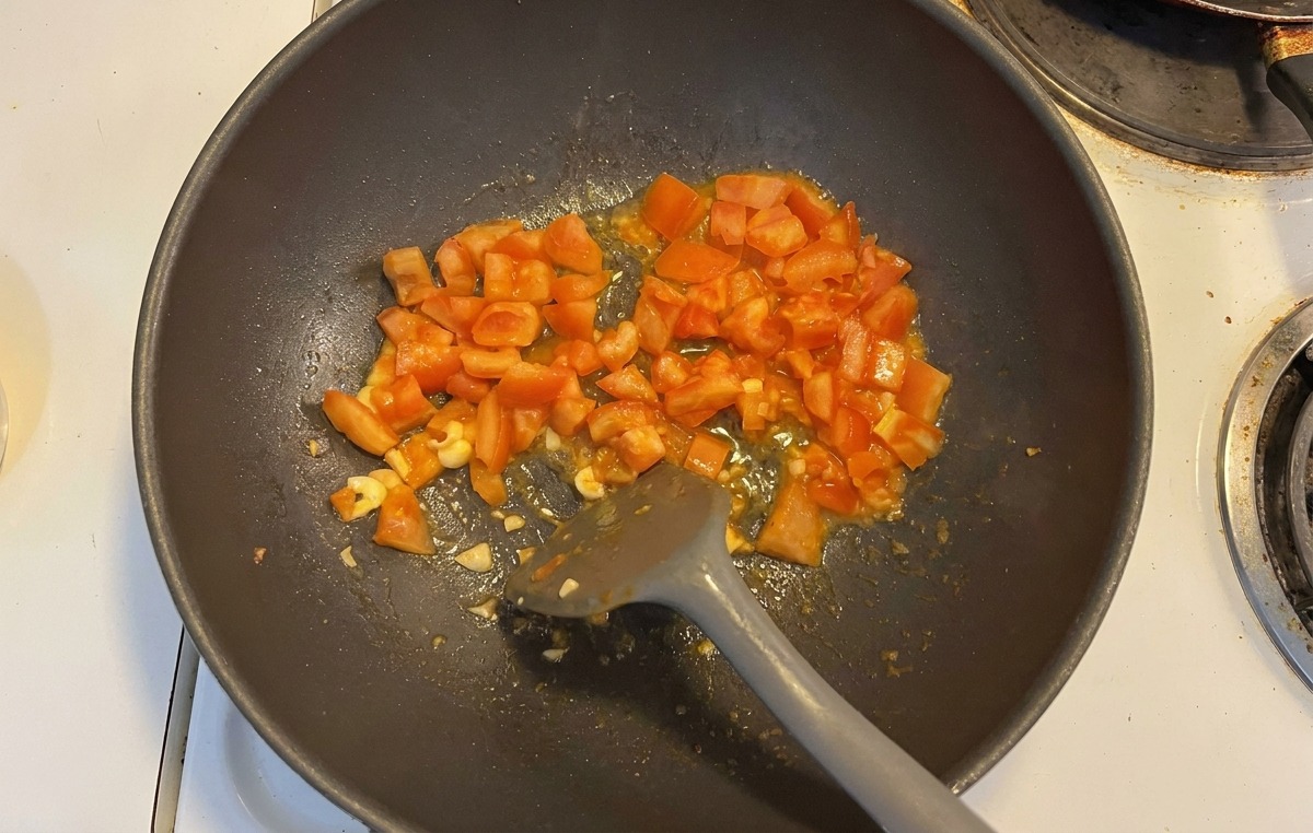Diced red tomatoes and sliced garlic being stir-fried with a gray spatula in a dark wok.