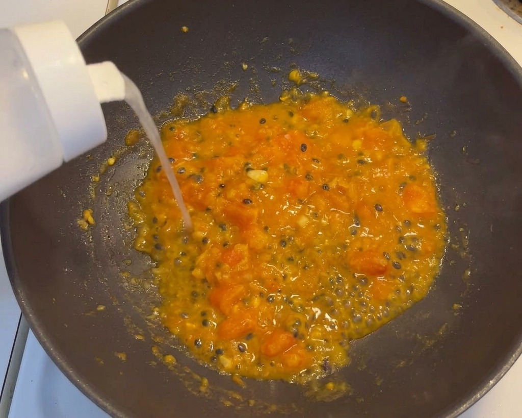 Shaking seasoning from a white dispenser into a bubbling wok of vibrant orange tomato and passion fruit soup base.