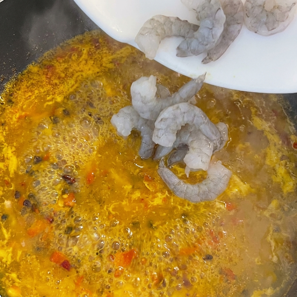 Raw, grey peeled shrimp sliding off a white plate into a pot of bubbling, bright yellow and orange broth.