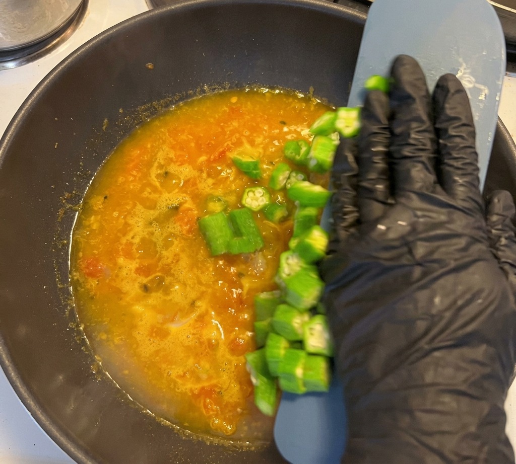 A gloved hand pushing sliced okra from a scraper into a bubbling yellow broth inside a dark pan.